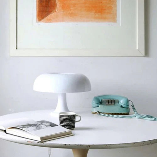 Weiße Tischlampe, Retro-Telefon, Kaffeetasse auf rundem Tisch vor Kunstwerk an weißer Wand.