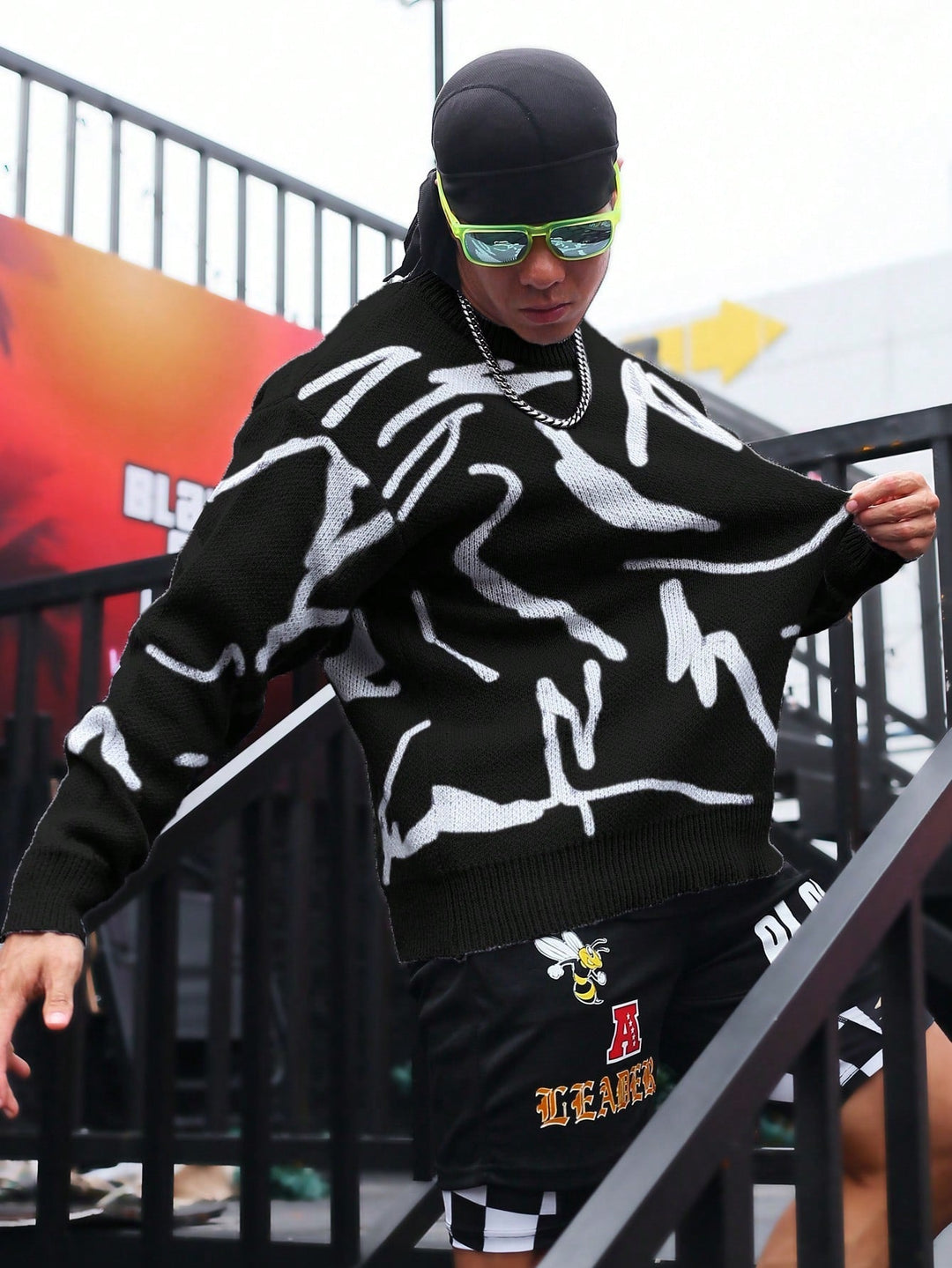 Mann in schwarzem Pullover mit weißem Muster, Sonnenbrille, schwarze Shorts, Treppe hinabsteigend.