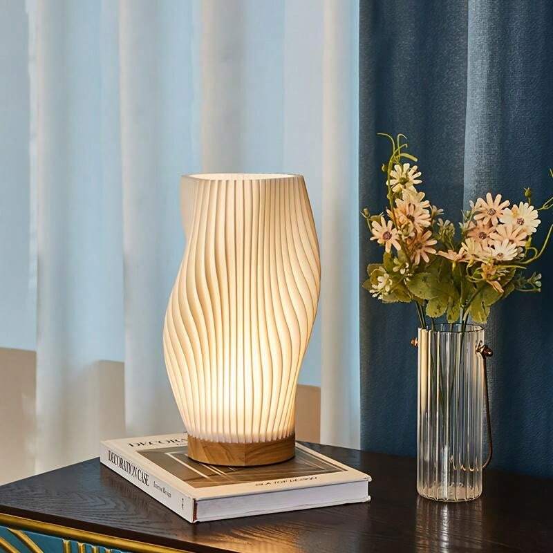 Moderne Tischlampe aus Holz mit wellenförmigem Design, warmes Licht, auf Tisch neben Blumen.