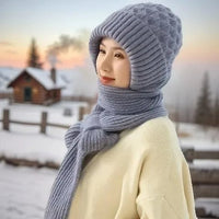 Frau mit grauem Strickmütze und Schal im Winter, vor schneebedeckter Landschaft und Holzhütte.