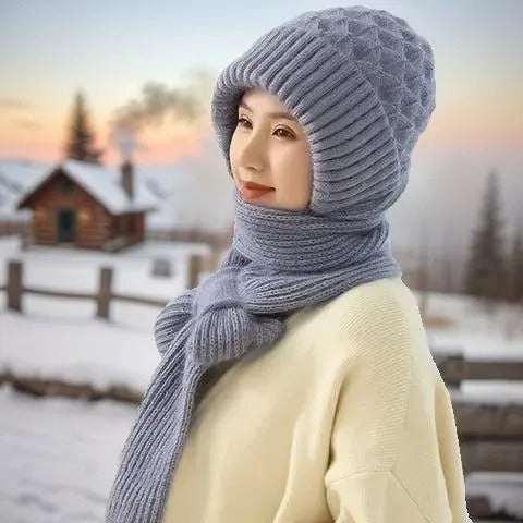 Frau mit grauem Strickmütze und Schal im Winter, vor schneebedeckter Landschaft und Holzhütte.