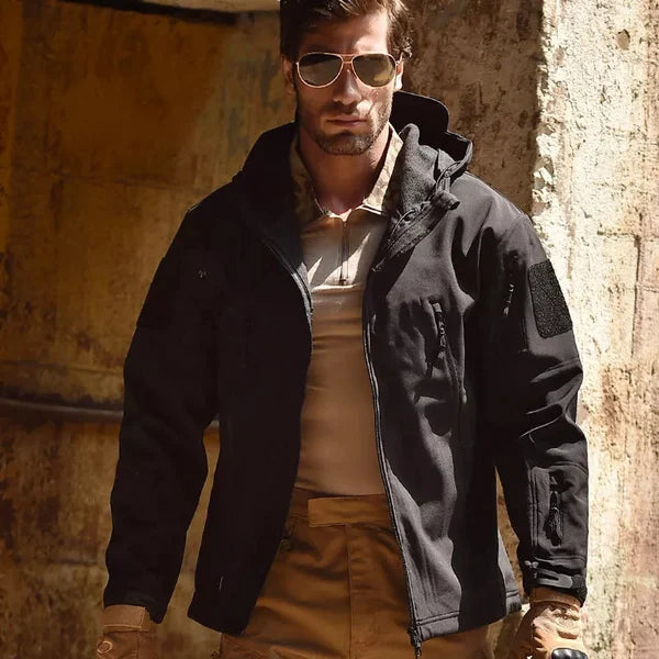 Mann in schwarzer taktischer Jacke mit Kapuze, Sonnenbrille, braune Hose, Outdoor-Kleidung.