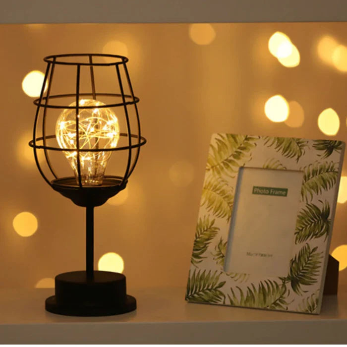 Schwarze Tischlampe mit Käfigdesign und warmem Licht neben Bilderrahmen mit Blattmuster.