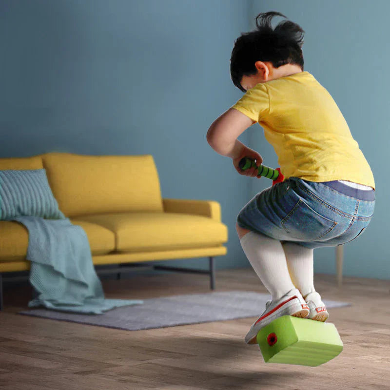 Kind spielt mit grünem Schaumstoff-Pogo-Stick im Wohnzimmer, gelbes Sofa im Hintergrund.