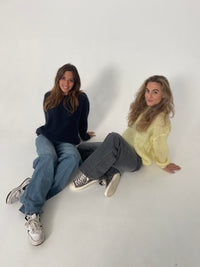 Zwei Frauen in Jeans und Pullovern, eine in Dunkelblau, die andere in Gelb, sitzen auf weißem Hintergrund.