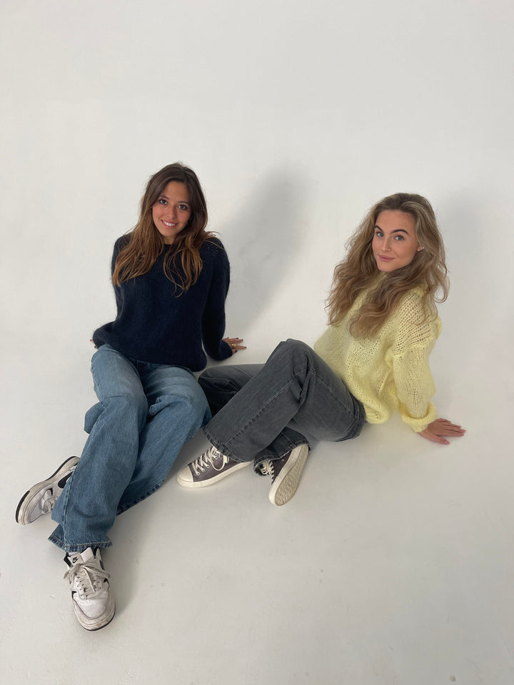 Zwei Frauen in Jeans und Pullovern, eine in Dunkelblau, die andere in Gelb, sitzen auf weißem Hintergrund.