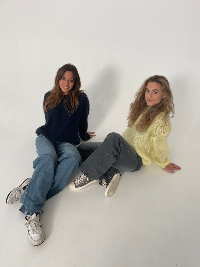 Zwei Frauen in Jeans und Pullovern, eine in Dunkelblau, die andere in Gelb, sitzen auf weißem Hintergrund.