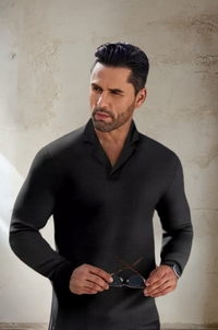 Mann in schwarzem Hemd hält Brille, vor rustikaler Wand, stilvolle Herrenmode.