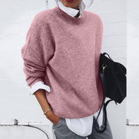 Frau in rosa Strickpullover, weißer Bluse, grauer Hose, hält schwarze Tasche.
