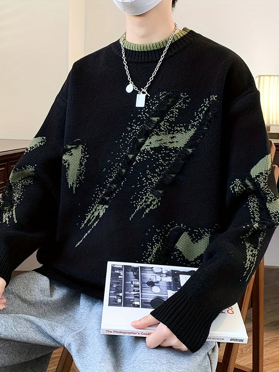 Mann in schwarzem Strickpullover mit grünem Muster, silberner Kette, hält ein Buch.