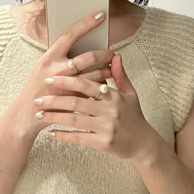 Frau mit Perlenring und Smartphone, beige Stricktop, elegante Maniküre, Nahaufnahme.