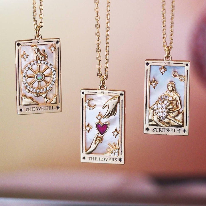Drei goldene Tarot-Anhänger mit Ketten, verziert mit Edelsteinen und Symbolen, auf rosa Hintergrund.