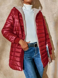 Frau in roter Daunenjacke, weißem Rollkragenpullover und blauer Jeans vor beiger Wand.