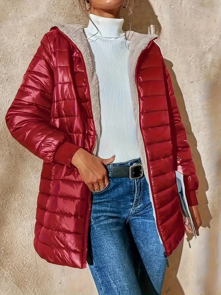 Frau in roter Daunenjacke, weißem Rollkragenpullover und blauer Jeans vor beiger Wand.
