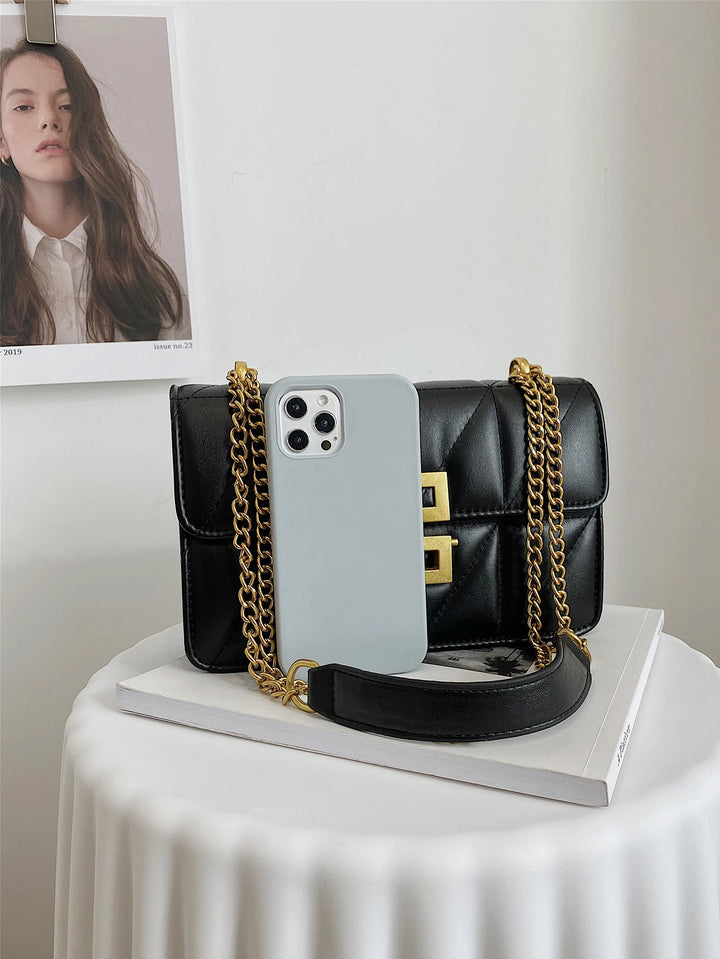 Schwarze Lederhandtasche mit goldener Kette und weißem Smartphone auf weißem Tisch.