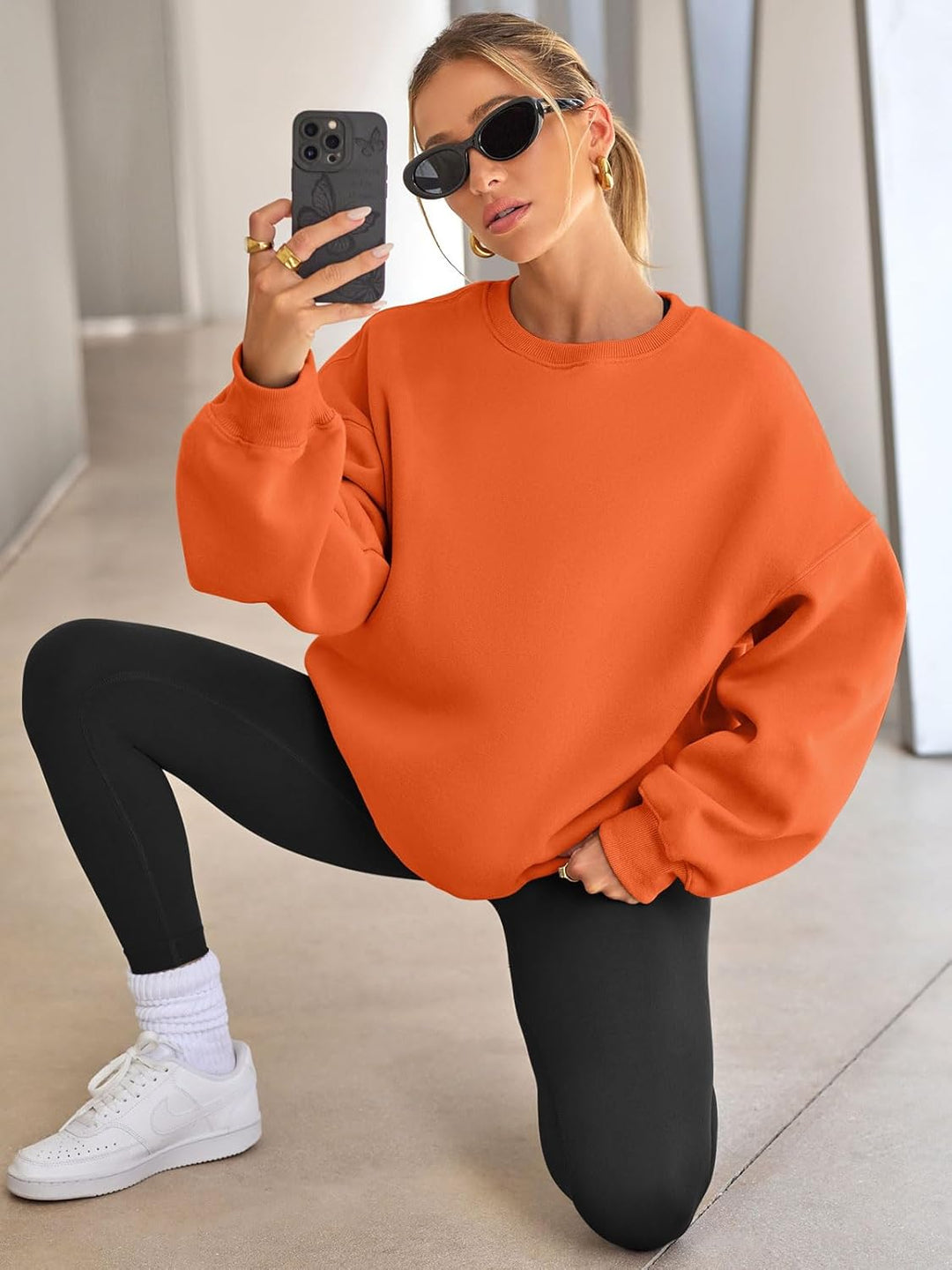 Frau in orangefarbenem Oversize-Pullover, schwarze Leggings, weiße Sneaker, Sonnenbrille, Smartphone.
