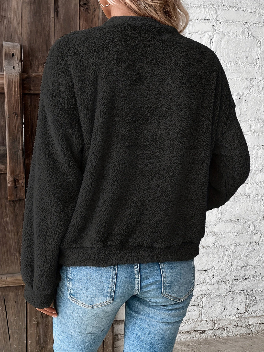 Frau in schwarzer Fleecejacke und blauer Jeans vor Holztür und weißer Wand.