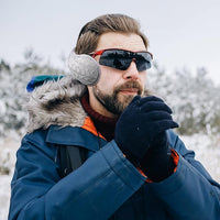 Mann mit blauer Winterjacke, Sonnenbrille und Ohrenschützer im verschneiten Wald.