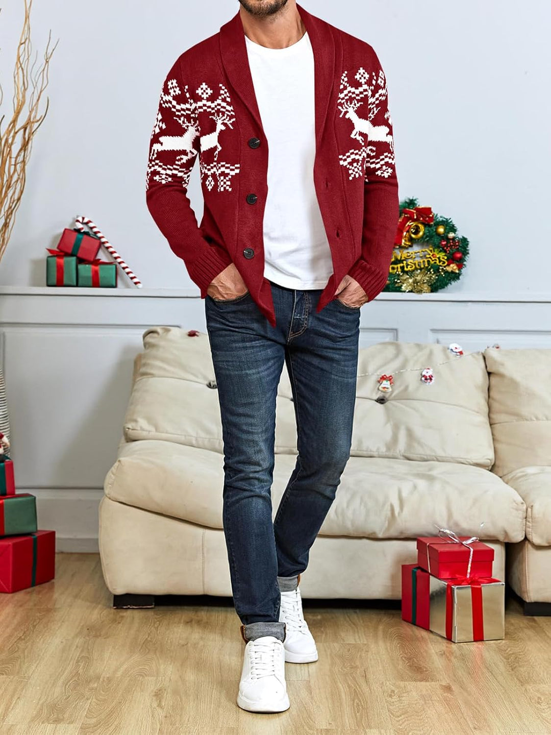 Mann in roter Strickjacke mit Rentiermotiv, weiße Sneakers, Jeans, Weihnachtsdeko im Hintergrund.