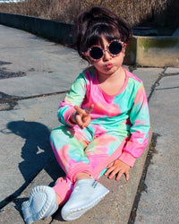 Mädchen in buntem Tie-Dye-Trainingsanzug und Sonnenbrille, sitzt auf Bürgersteig.