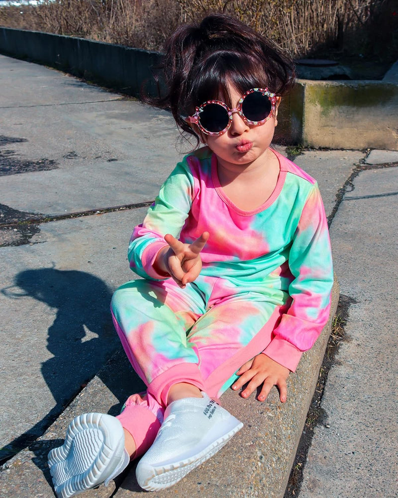 Mädchen in buntem Tie-Dye-Trainingsanzug und Sonnenbrille, sitzt auf Bürgersteig.