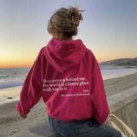Frau am Strand in pinkem Hoodie mit Aufschrift, Sonnenuntergang, Meereshintergrund, Freizeitkleidung.