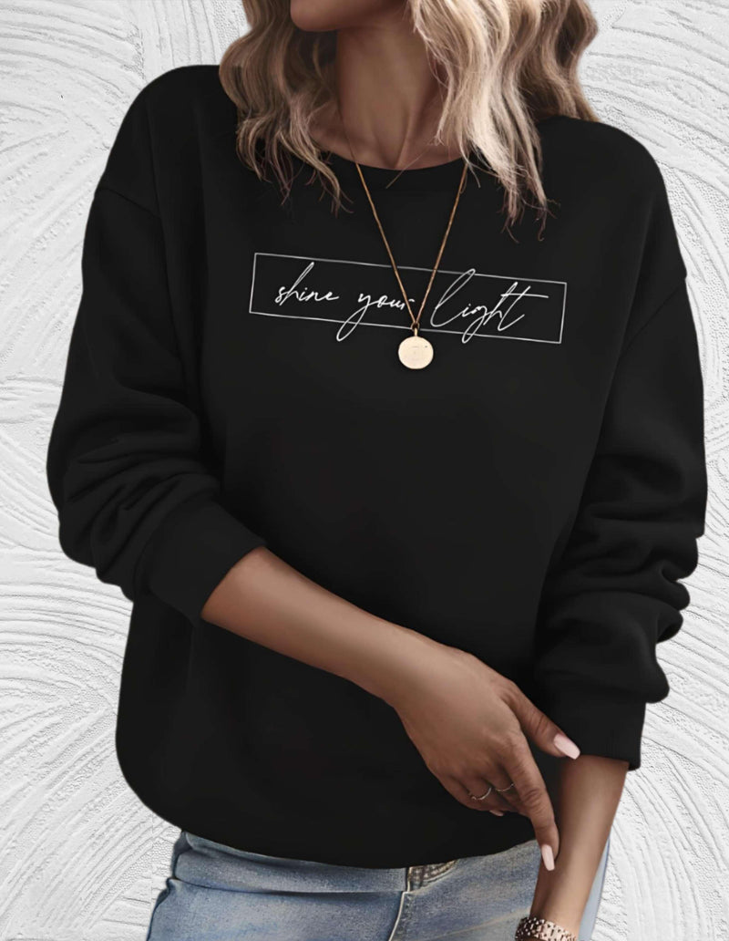 Frau in schwarzem Sweatshirt mit "shine your light" Aufdruck, goldene Halskette, lässiger Stil.