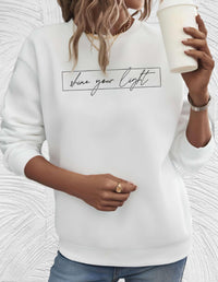 Frau in weißem Sweatshirt mit "shine your light"-Aufdruck, hält weißen Becher.