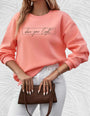 Frau in rosa Sweatshirt mit "shine your light"-Aufdruck, weiße Hose, braune Handtasche.