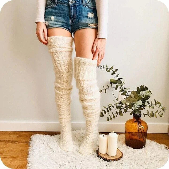 Frau trägt hohe, cremefarbene Stricksocken aus Wolle, kombiniert mit Jeansshorts, auf Teppich.