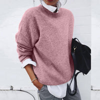Frau in rosa Strickpullover, weißer Bluse, grauer Hose, hält schwarze Tasche.