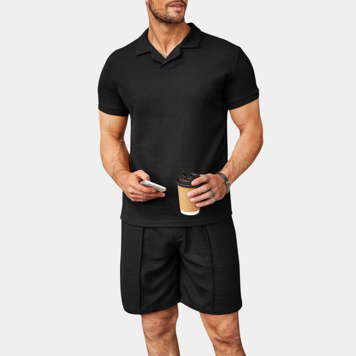 Mann in schwarzem Polohemd und Shorts, hält Kaffeebecher und Smartphone.