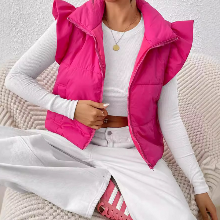 Frau in pinker Steppweste, weißem Langarmshirt, weißen Hosen und rosa Sneakers auf Sofa.