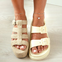 Zwei verschiedene Sandalen an Füßen, eine aus geflochtenem Stroh, die andere aus cremefarbenem Kunststoff.