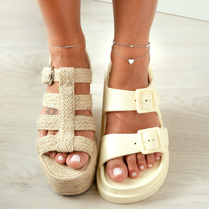 Zwei verschiedene Sandalen an Füßen, eine aus geflochtenem Stroh, die andere aus cremefarbenem Kunststoff.