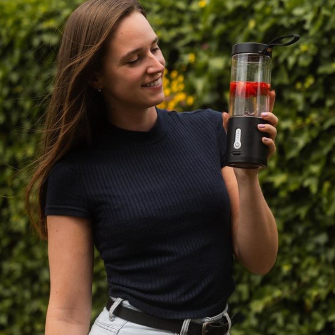 Frau mit tragbarem Smoothie-Maker, schwarz, mit Erdbeeren, im Freien, vor grünem Hintergrund.