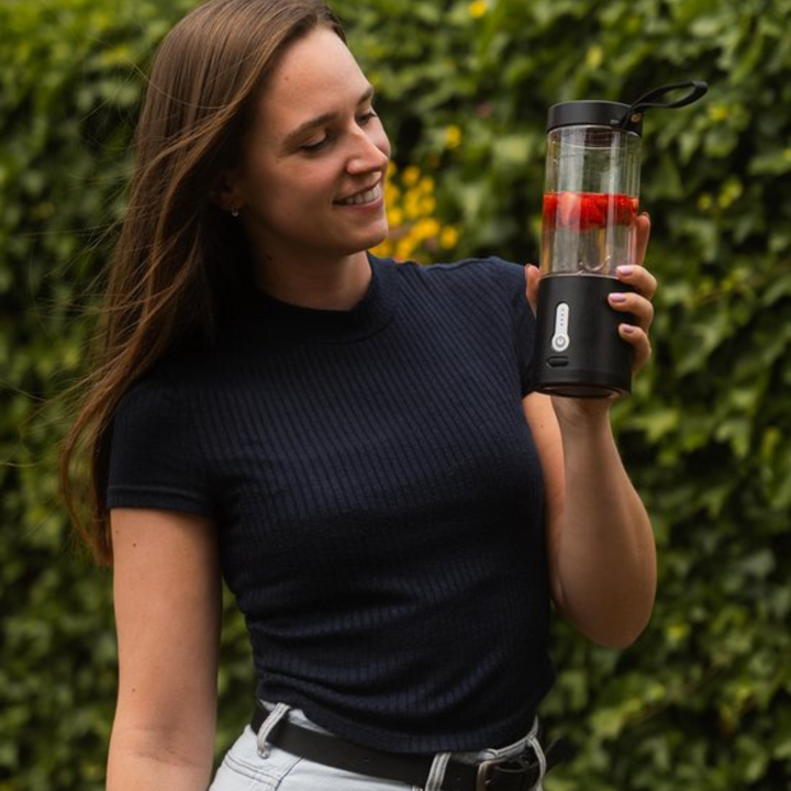 Frau mit tragbarem Smoothie-Maker, schwarz, mit Erdbeeren, im Freien, vor grünem Hintergrund.