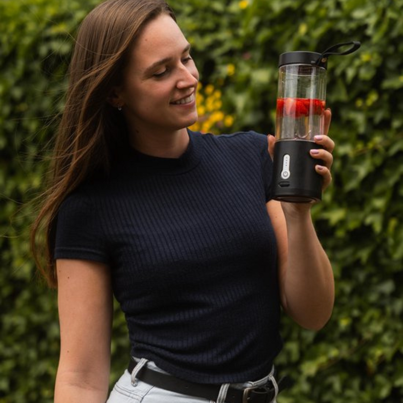Frau mit tragbarem Smoothie-Maker, schwarz, mit Erdbeeren, im Freien, vor grünem Hintergrund.