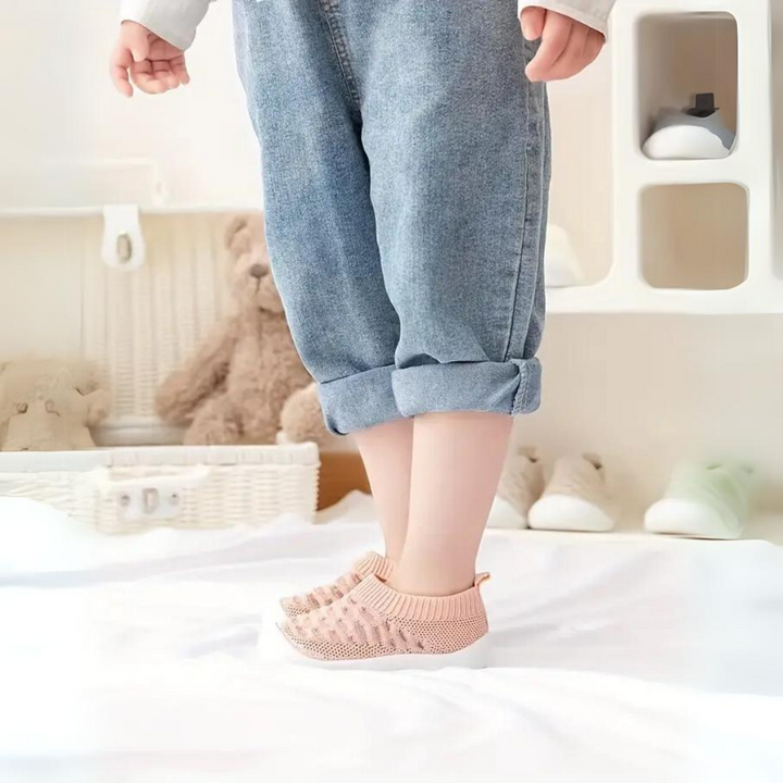 Kleinkind trägt rosa Strickschuhe, blaue Jeans, steht auf weißem Boden, Kinderzimmer im Hintergrund.