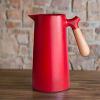Rote Thermoskanne mit Holzgriff, skandinavisches Design, auf Holztisch vor Ziegelwand.