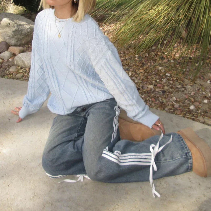 Frau in blauer Strickpullover und weiten Jeans mit Seitenstreifen, kniend im Freien.