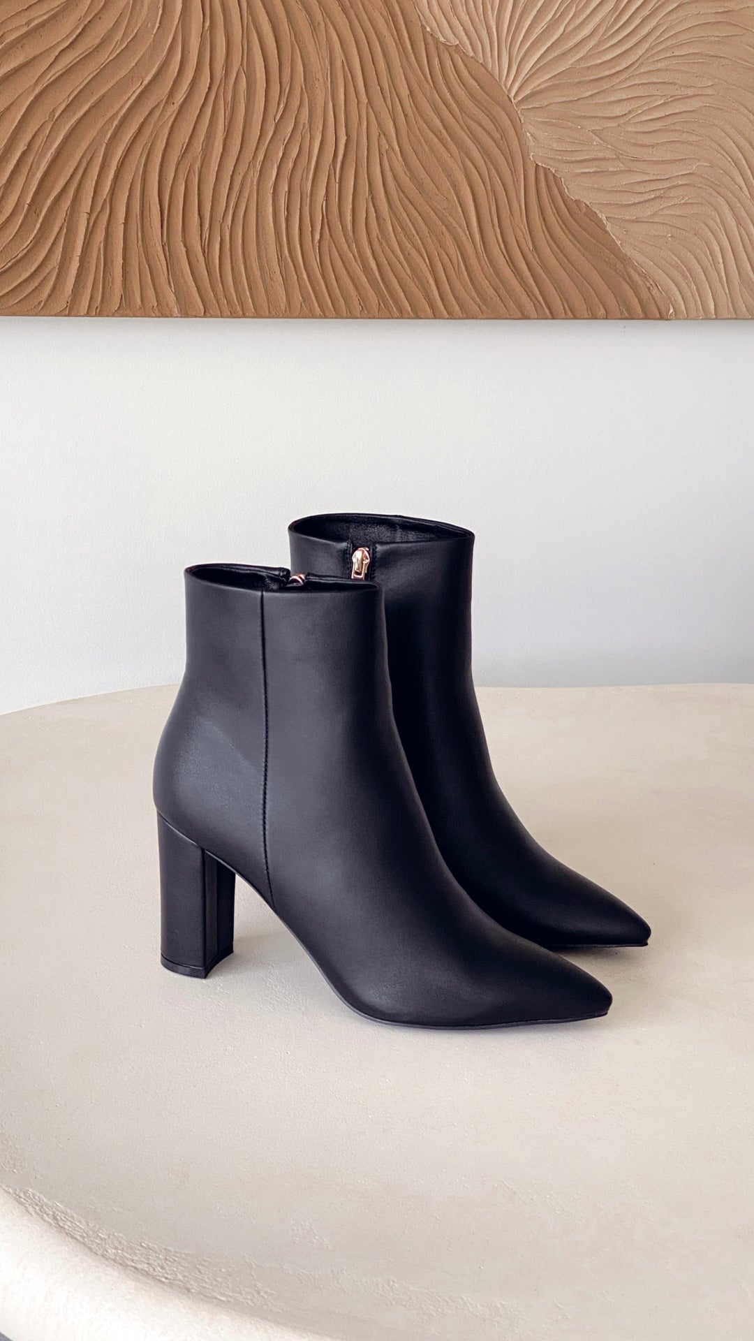 Schwarze Damenstiefeletten aus Leder mit Blockabsatz auf beigem Tisch vor Wandkunst.