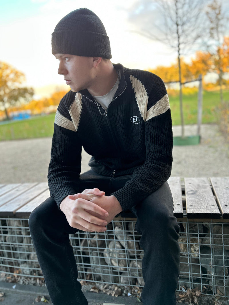 Mann in schwarzer Strickjacke mit Mütze, sitzt auf Bank im Herbstpark.