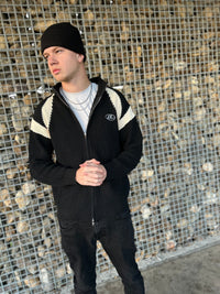 Mann in schwarzer Strickjacke mit weißen Streifen und Mütze vor Steinwand.