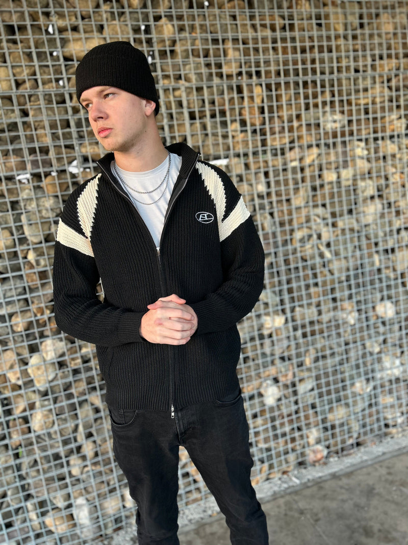 Mann in schwarzer Strickjacke mit weißen Streifen und Mütze vor Steinwand.