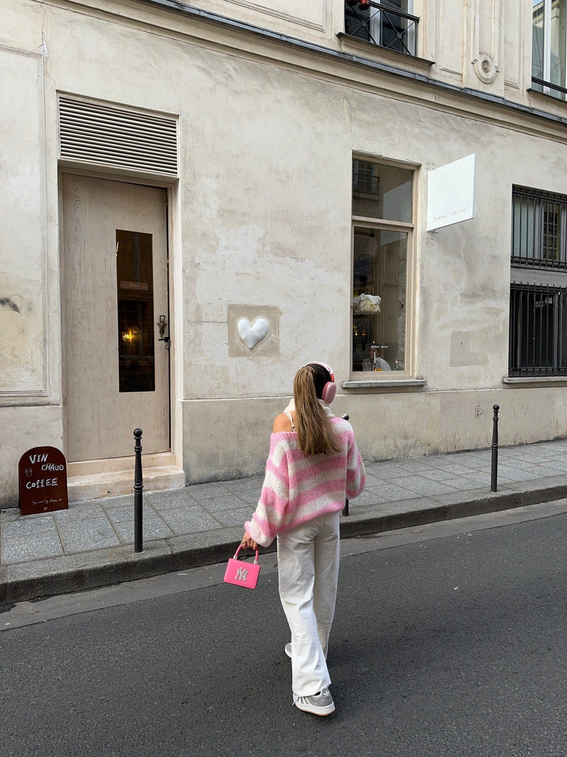 Frau in rosa-weißem Pullover und weißer Hose, trägt rosa Tasche, geht auf Pariser Straße.