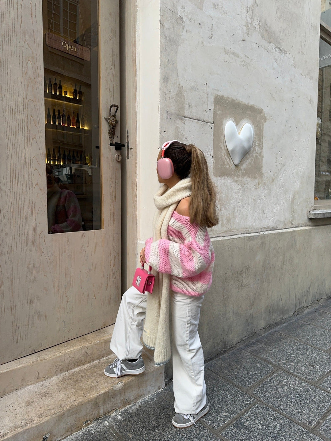 Mädchen in rosa-weißem Pullover, weißer Hose, mit Kopfhörern und Tasche vor Geschäftstür.