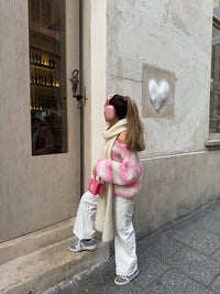 Mädchen in rosa-weißem Pullover, weißer Hose, mit Kopfhörern und Tasche vor Geschäftstür.