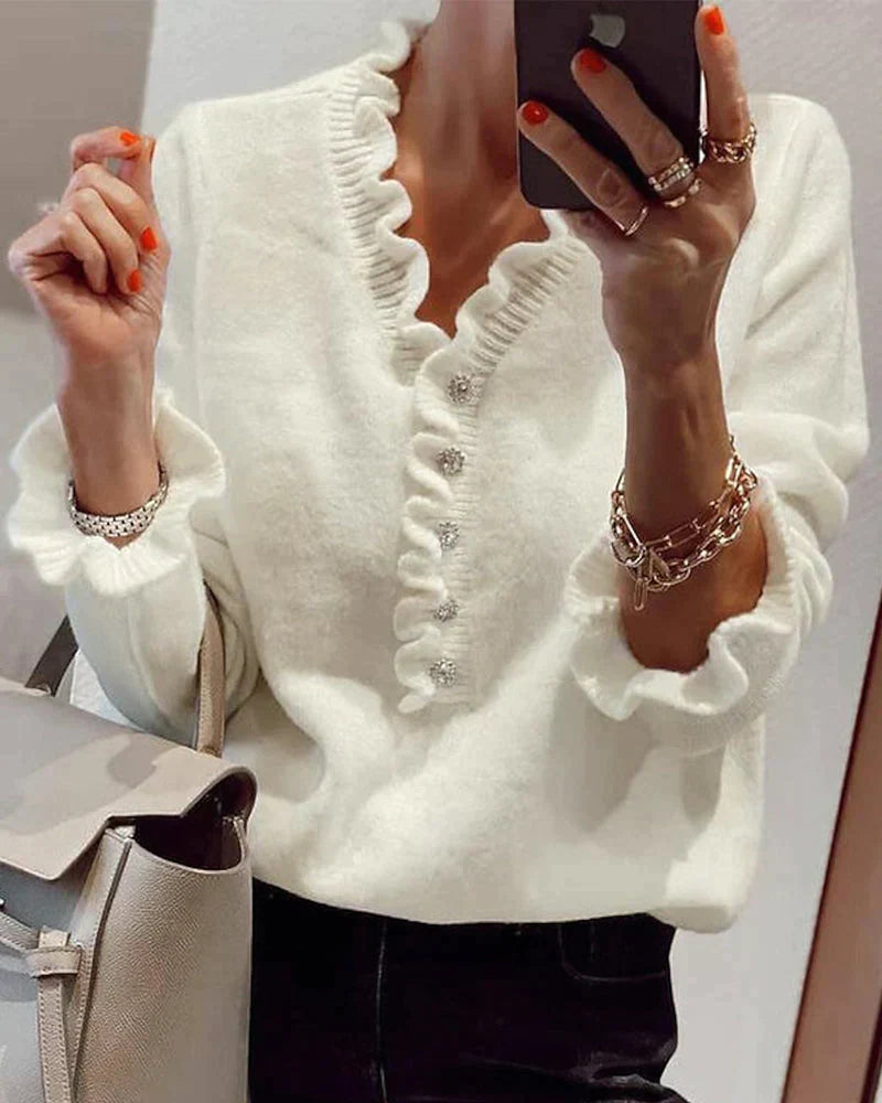 Frau in weißem Rüschenpullover mit Knöpfen, hält Smartphone, trägt Schmuck, modisch elegant.