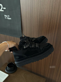 Schwarze Sneaker mit dicker Sohle, Fellfutter und gemusterten Schnürsenkeln vor Holzwand.